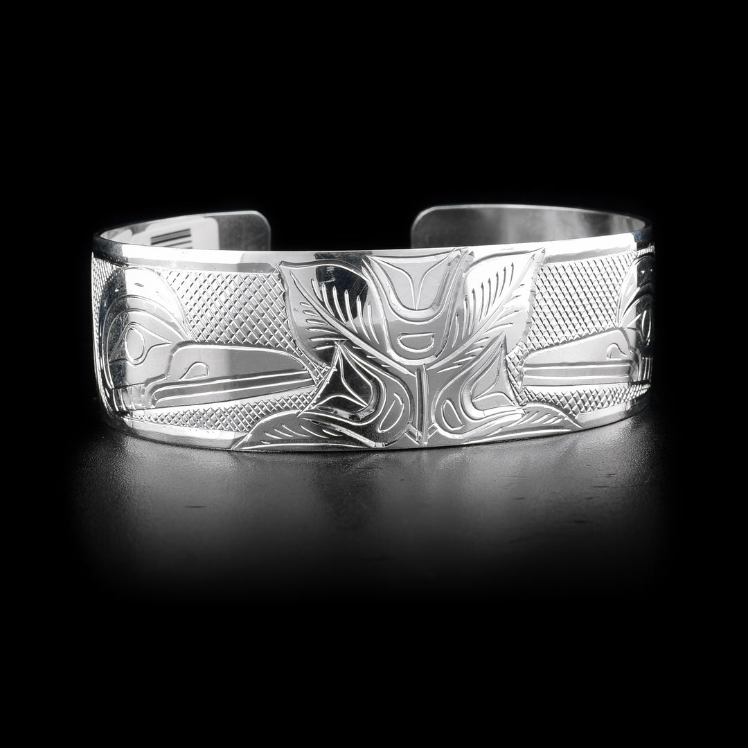 Bracelet has mirroring effect. Two flowers in the center with leaves around. At each flower there is a hummingbird. Cross-hatching background. Hand-carved by Kwakwaka’wakw artist William Cook.