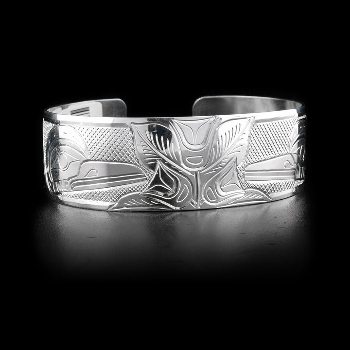 Bracelet has mirroring effect. Two flowers in the center with leaves around. At each flower there is a hummingbird. Cross-hatching background. Hand-carved by Kwakwaka’wakw artist William Cook.