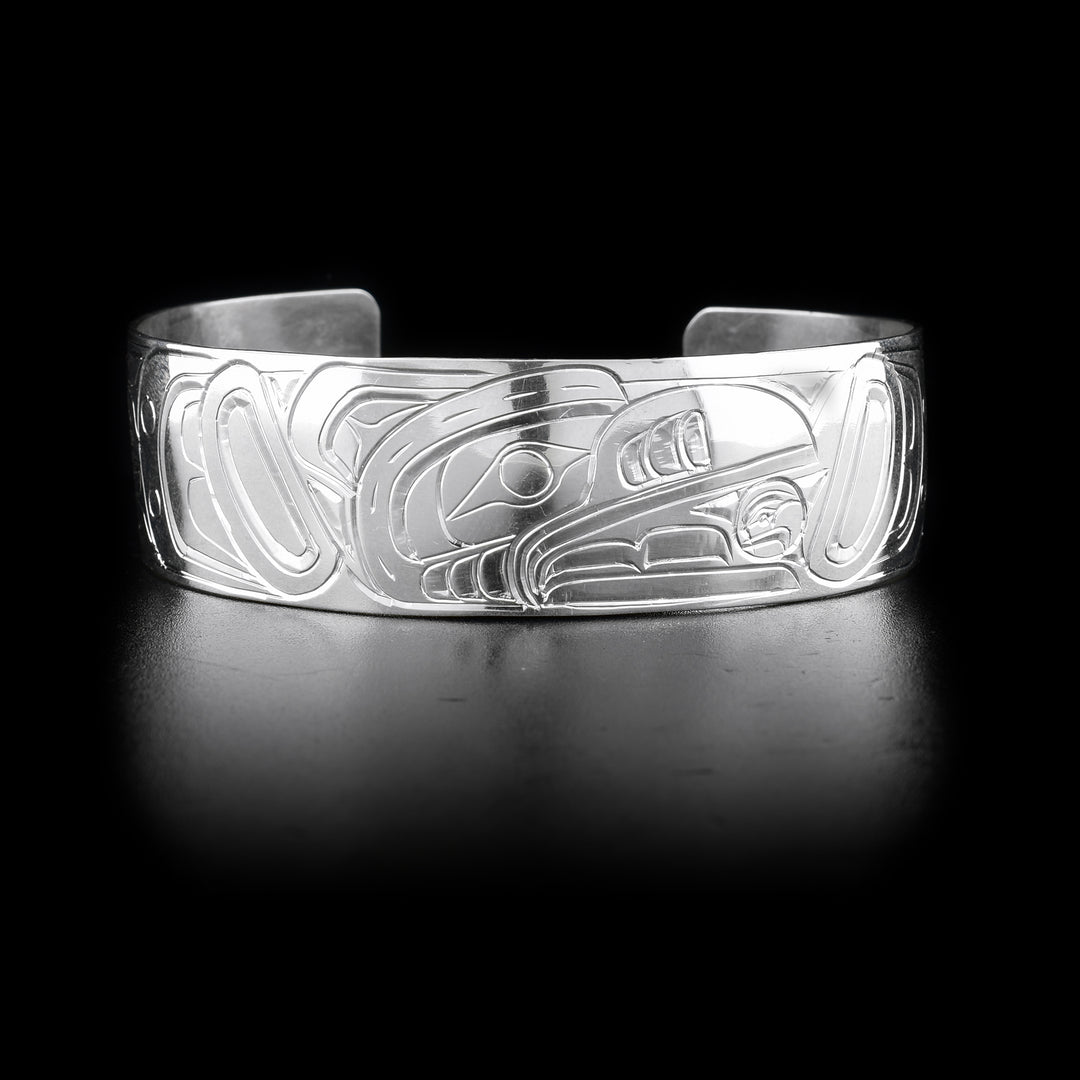 This Raven and Sun bracelet has a wide and sterling silver band with a gap in the back. There are carvings across the surface of the band depicting the Raven stealing the Sun. The scene depicts an indigenous legend.