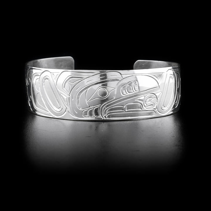 This Raven and Sun bracelet has a wide and sterling silver band with a gap in the back. There are carvings across the surface of the band depicting the Raven stealing the Sun. The scene depicts an indigenous legend.