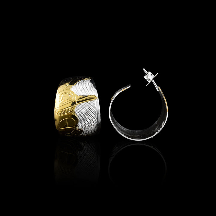 These Hummingbird hoop earrings are made from sterling silver and 14K gold. They have wide hoop stud that taper slightly on both ends. There is a side profile depiction of the hummingbird caved into the hoop using the gold. Its wing is at its side and its head is looking side ways. There are cross hatching designs made in the background of the scene.