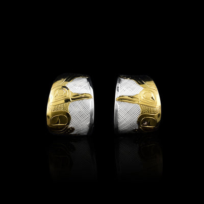 These Hummingbird hoop earrings are made from sterling silver and 14K gold. They have wide hoop stud that taper slightly on both ends. There is a side profile depiction of the hummingbird caved into the hoop using the gold. Its wing is at its side and its head is looking side ways. There are cross hatching designs made in the background of the scene.