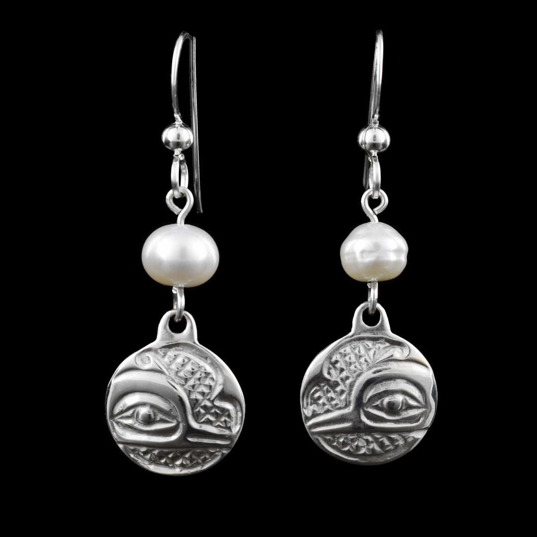 The hummingbird earrings are made of silver with a white pearl accent. The earrings have a round silver charm at the bottom, engraved with a hummingbird in traditional indigenous style. Above each charm is a single white pearl, adding an elegant contrast to the detailed silver design. The earrings are attached to fishhook-style ear wires, which make them the perfect silver and pearl dangle earrings.