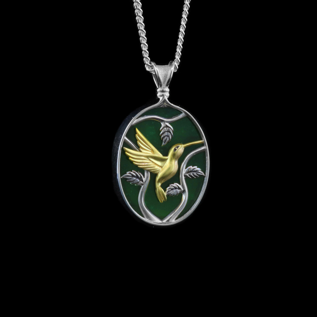 This jade pendant necklace has an oval shaped with a depiction of a Hummingbird flying around a tree. The hummingbird is made from 14K gold and the jade and three designs are set in sterling silver.