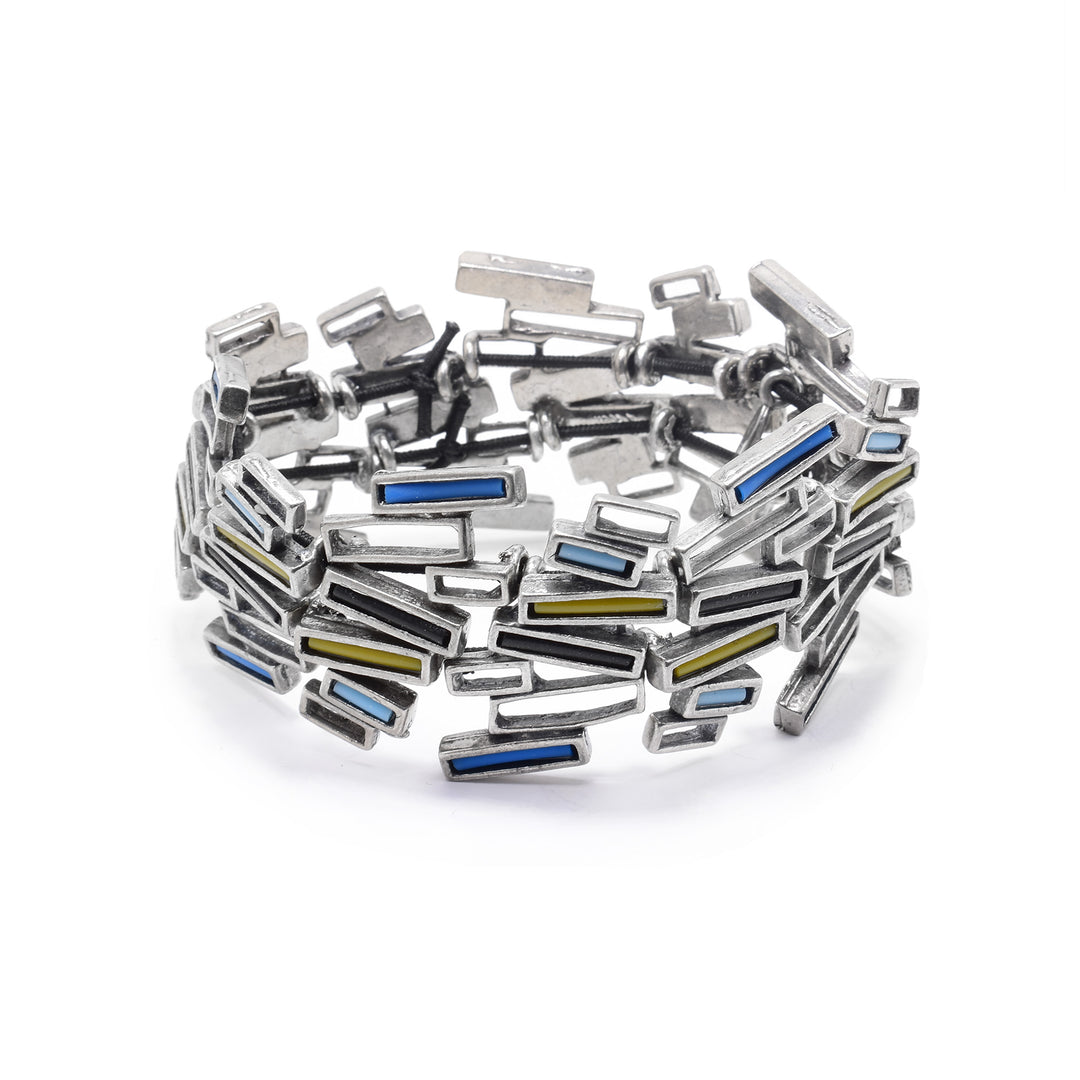 This pewter cuff bracelet is made using a combination of small, pewter rectangles that have their middles cutout. The rectangles are attached together to form this bangle with a solid, all around band. The band is wide and have a rugged texture due to the angles of the rectangles. There are black, yellow, blue and turquoise colored glass pieces placed in the center of some of the rectangles around the piece.