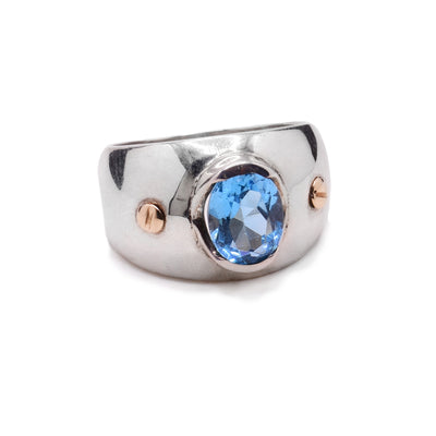 These blue topaz rings have wide, tapered and sterling silver bands with a large oval shaped blue topaz gem embossed in the center of the solitaire ring. There are two 14K gold studs placed on either side of the gem.