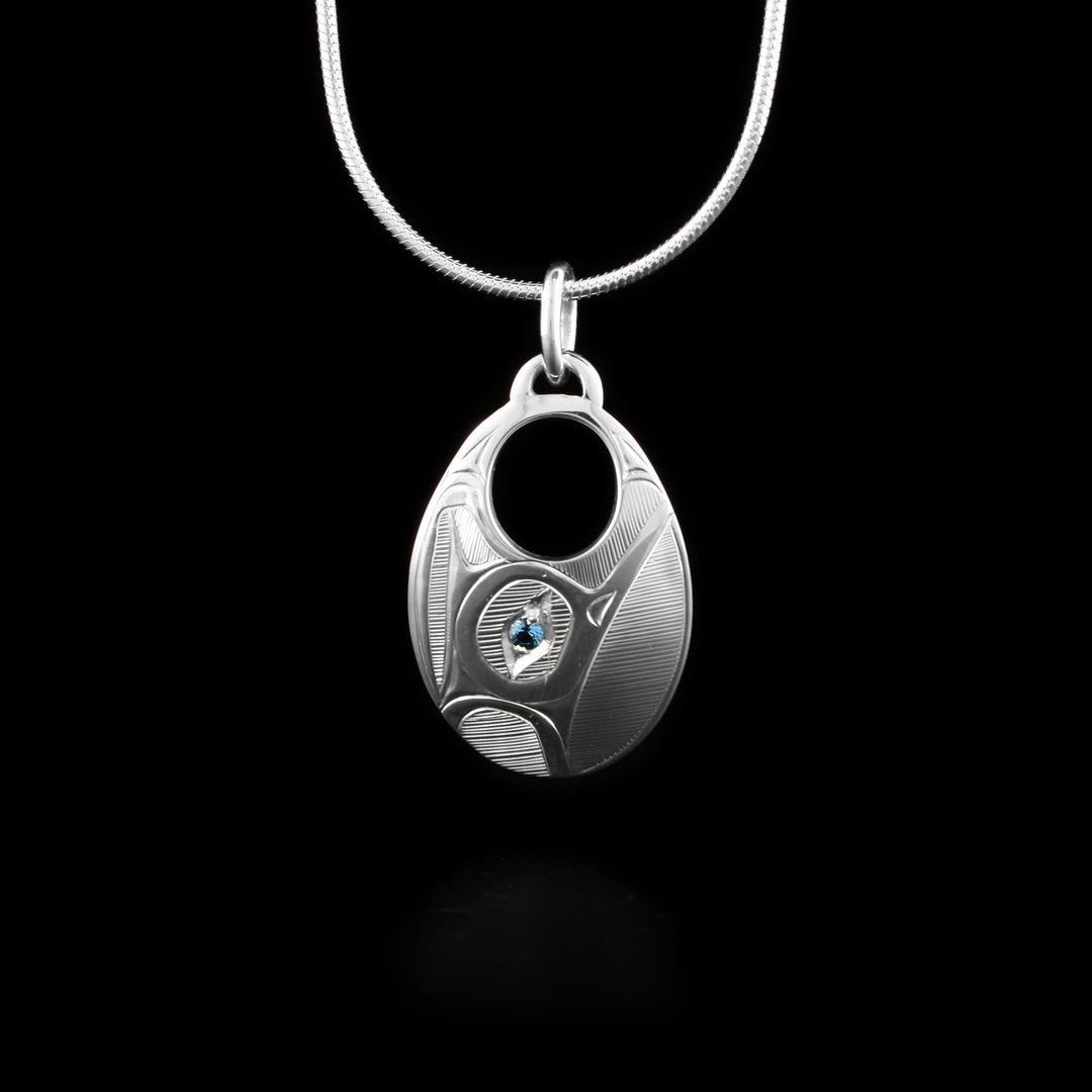 This Hummingbird pendant is wide, oval shaped and has a circle cutout on the top of the pendant. The Hummingbird legend is depicted in side profile and its head is looking upwards. In the spaces behind the legend, there are hatching patterns. There is a blue topaz gems imbedded in the eye of the Hummingbird.