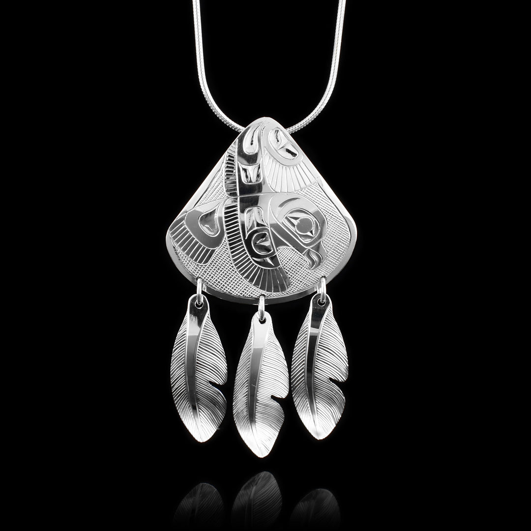 This Eagle feather pendant is made from sterling silver and has a wide, rounded teardrop shape. There is a side profile depiction of the Eagle carved into it with both its wings outstretched. The eagle appears to be in flight and there are feather designs included on the wings and tail. There are three, sterling silver feathers dangling off of this pendant on jump rings.