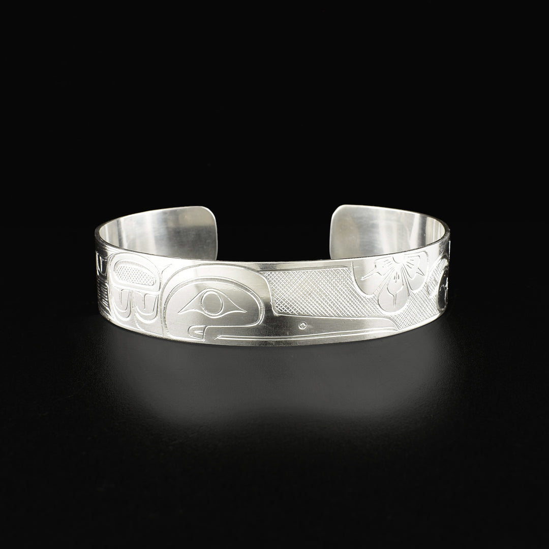 Detailed cuff bracelet depicting a hummingbird in a floral setting. Side-view of the hummingbird’s face is in the center. Design is made from cross-hatching, ovoids and lines.