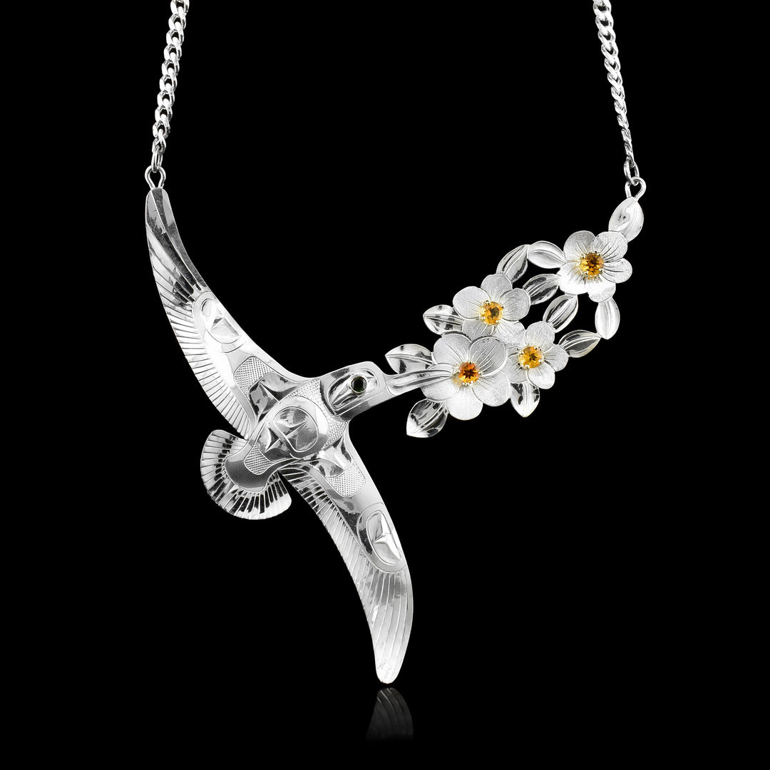 This exquisite sterling silver hummingbird necklace is an authentic creation by Tlingit artist Fred Myra. The design depicts a singular formline hummingbird journeying toward a branch of wild roses. Bringing vibrant color to the piece, the blossoms feature radiant citrine centers set in 14k gold prongs, complemented by a rich, dark green tourmaline gemstone for the hummingbird’s eye.