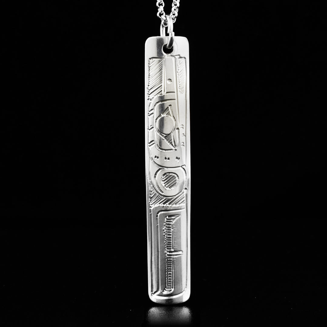 This Hummingbird pendant is made out of sterling silver. It has the shape of a long rectangle. The face of the Hummingbird is carved on it, facing upwards.