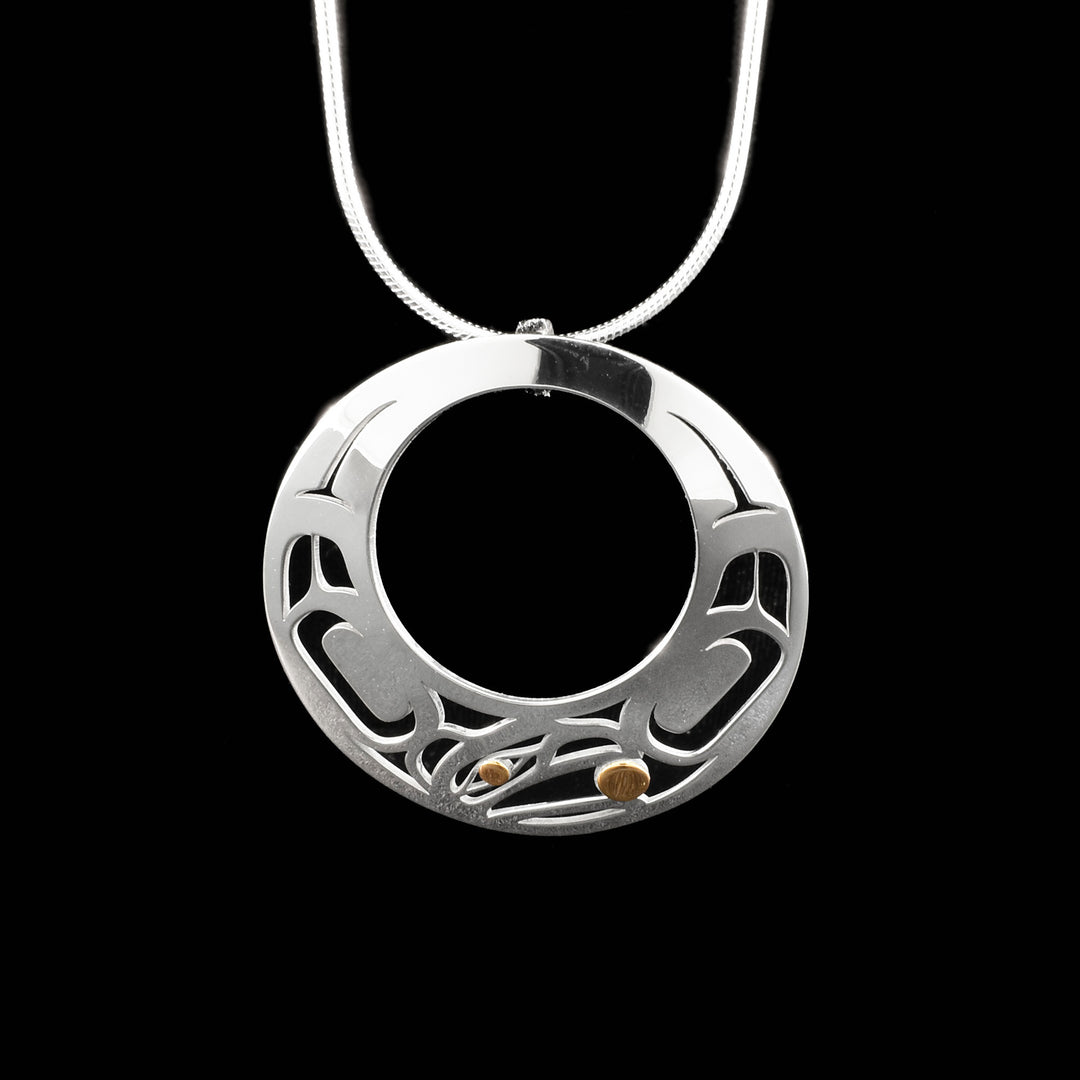 Round sterling silver cut-out pendant with pierced raven design. Raven’s head on bottom, facing left, with ball in beak. Designs on either side of the head depict wings. 18K gold in eye and ball. By Tahltan artist Grant Pauls.