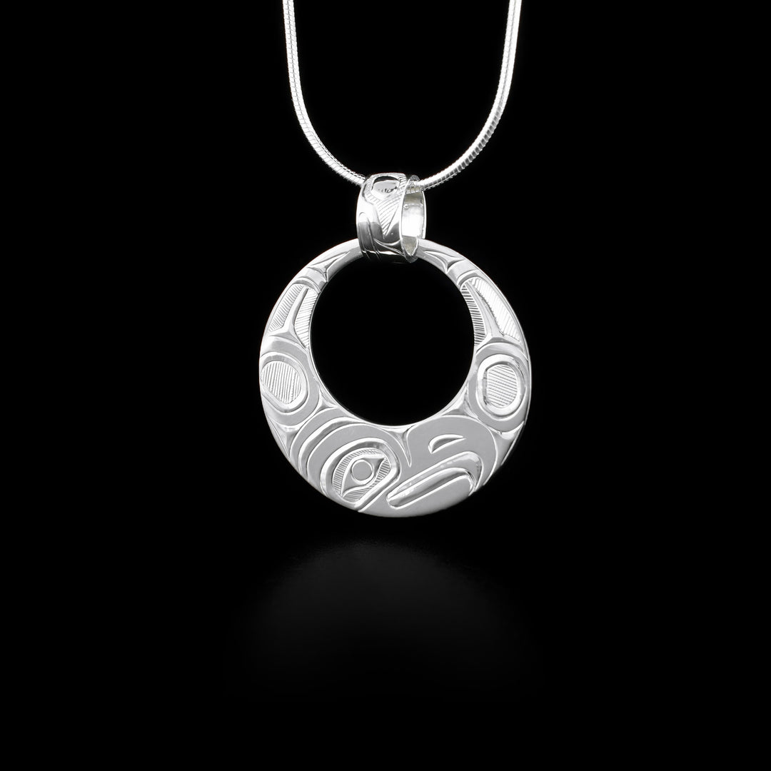 This Eagle pendant is made from sterling silver and has a circle shape with a large, circle shaped cutout in the top. The head of the Eagle is depicted at the side view and its at the bottom of the pendant. There are wing designs extending up on either side carved into the pendant. The pendant hangs from a decorated, wide bail that has designs carved into it.