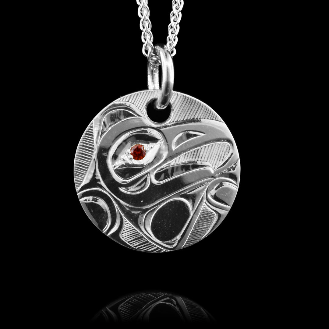 This Sterling Silver Eagle Pendant with Garnet is hand carved by artist, Hollie Bartlett. The piece is a sterling silver circle and the Eagle is depicted in side view on it. The head of the eagle is in the center and its wing is bent around it at the bottom of the piece. There is a red, garnet gem embedded in the eye of the legend.