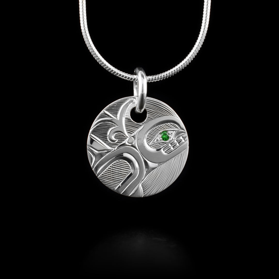 This Orca whale pendant is made from sterling silver and has a small, circle shape. The Orca is carved into the piece depicted in a side view. The upper half of the Orca is shown and there is a spout of water coming from its blow hole. Behind the Orca, there are hatching designs. There is a green tsavorite gem held in the eye of the legend.
