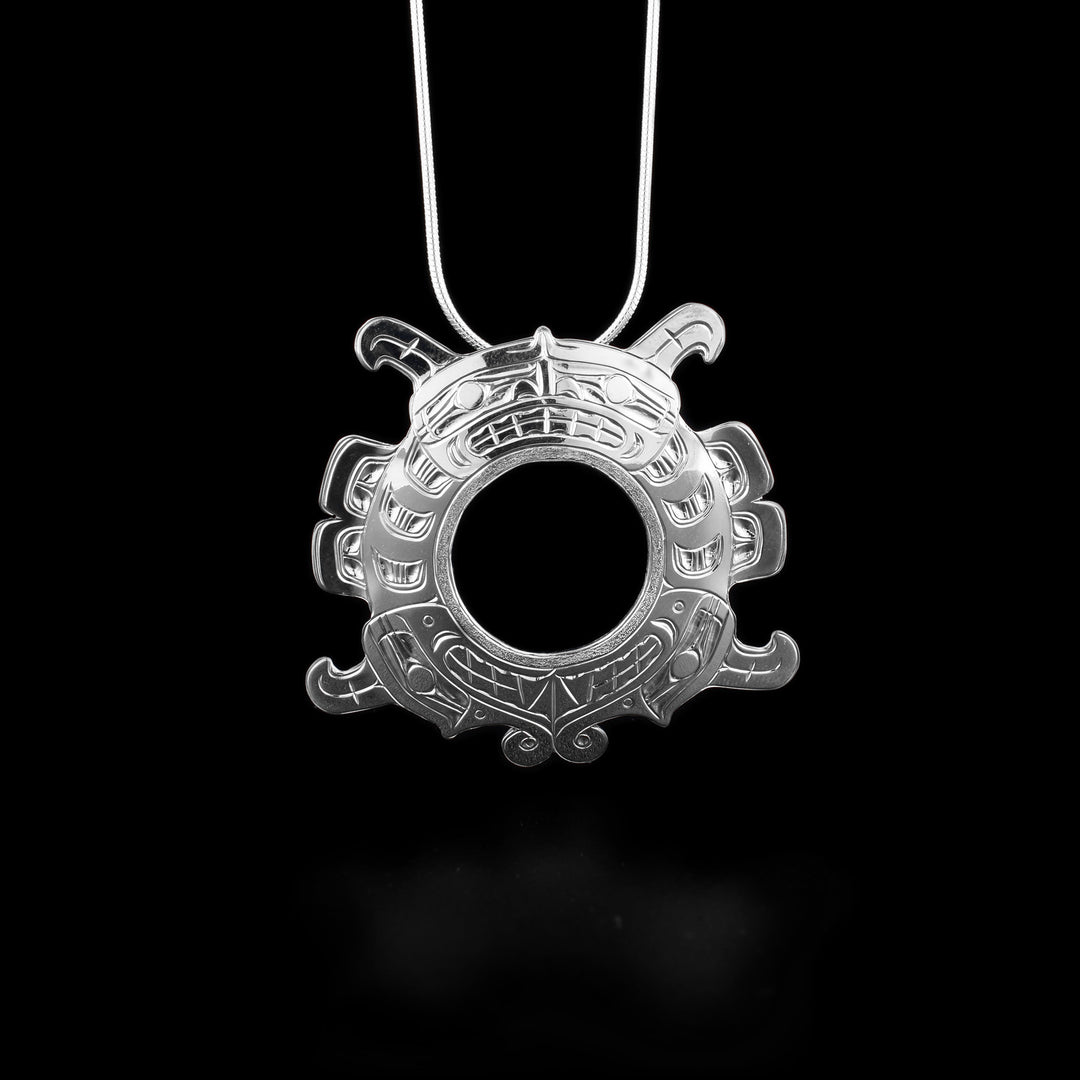 Sisiutl curls around into ring shape. Two serpent heads meet at the bottom and a humanoid face in center of serpent is at the top. Sisiutl has been cut out of silver, giving the edges an almost graphic look. Textured border on underside of Sisiutl makes the central hole circular. Hidden bail on back.