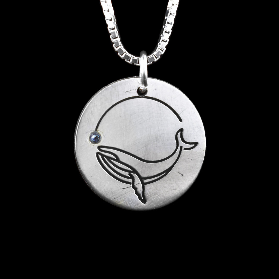 Sterling Silver Whale Pendant with Gemstone. A dainty circular pendant with an etched blue whale curved upwards towards a blue gem stone.