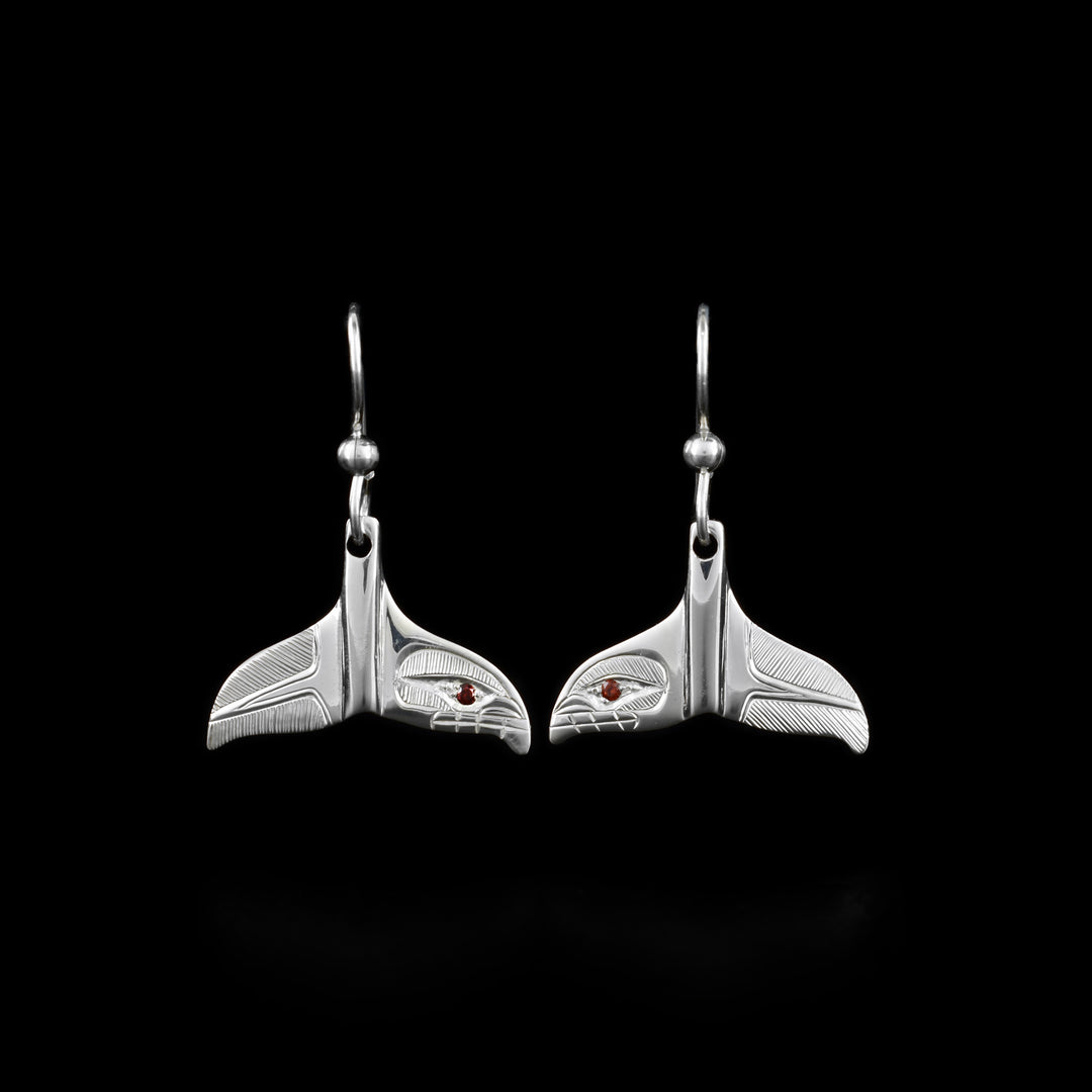 These Orca whale earrings are made from sterling silver and have whale tail shaped hangs. On one of the fins, there are carvings depicting the head of the Orca and on the other, there are fine, line patterns depicting the tail. There are garnet gems embedded in the yes of the Orcas.