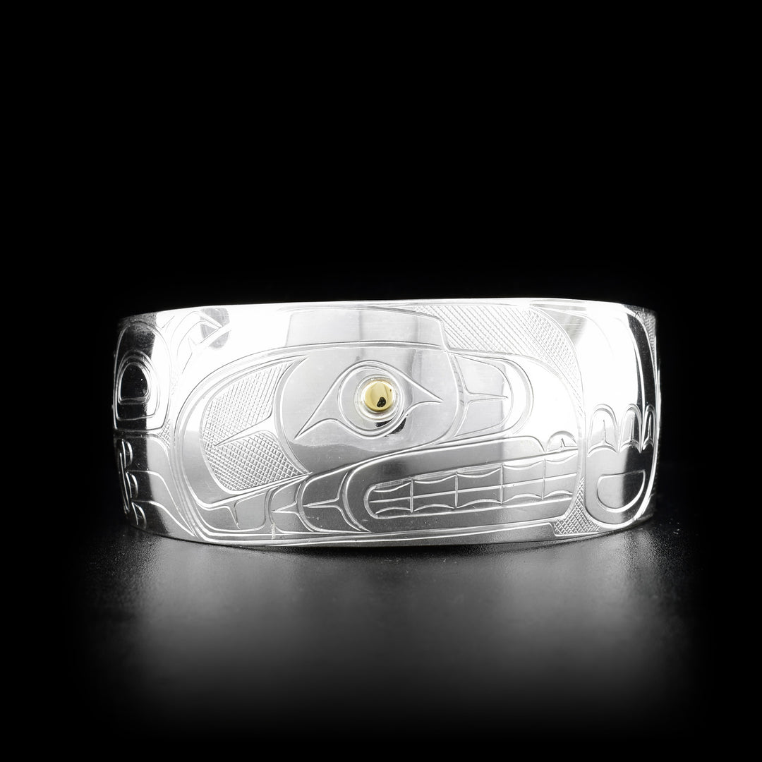 This silver and gold bracelet is made from sterling silver and 14K gold and has a wide band that tapers slightly. There is a depiction of the Bear carved into the surface of the band and the eye of the Bear is made from 14K gold.