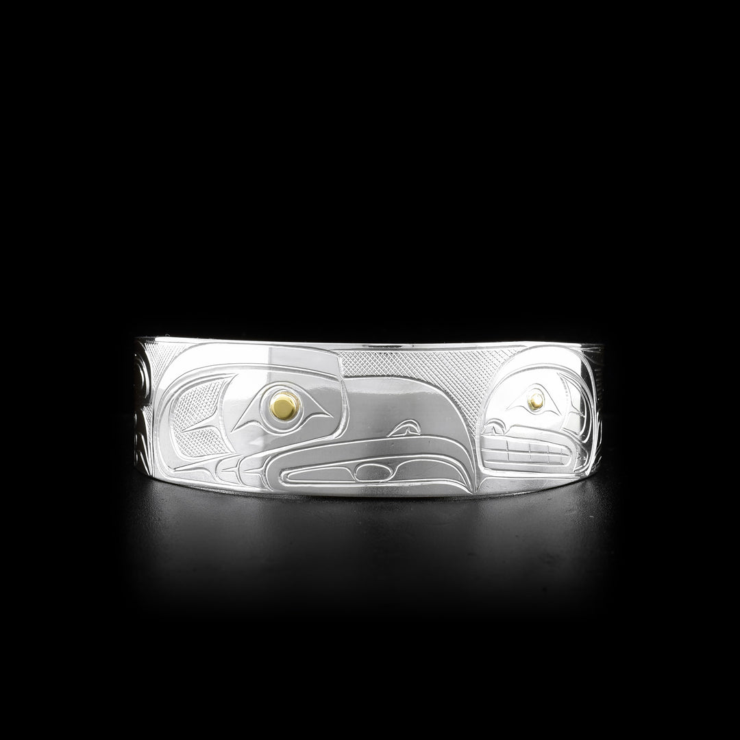 This silver and gold bracelet is made from sterling silver and 14K gold and has a wide band with a gap in the back. There are depictions of the Raven and the Orca carved into the surface of the band. The eyes of the legends are made from 14K gold.