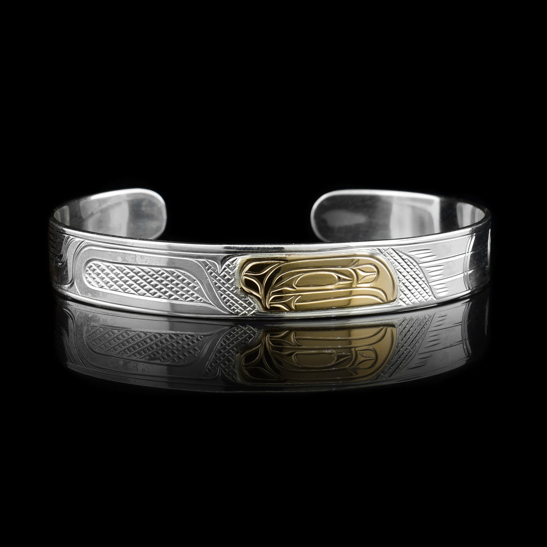 This sterling silver cuff bracelet has a single, thin band with a gap in the back. There are carvings on the surface of the band depicting the Raven and the head of it is made from 14K gold. 