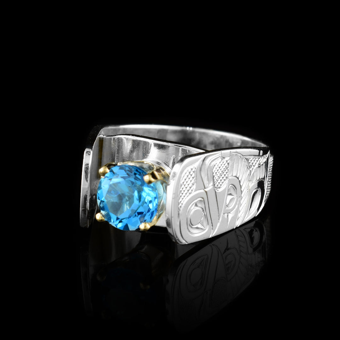 This blue topaz solitaire ring has a wide and tapered band that extends in cathedral style above the blue topaz gem that is set in 14K gold claws. There are carvings made on the band that depict the Eagle. The Eagle is facing side ways at a side profile and has its wing extended out with feather details. Its eye, beak and tail are detailed and there are cross hatching designs in the background of the Scene.