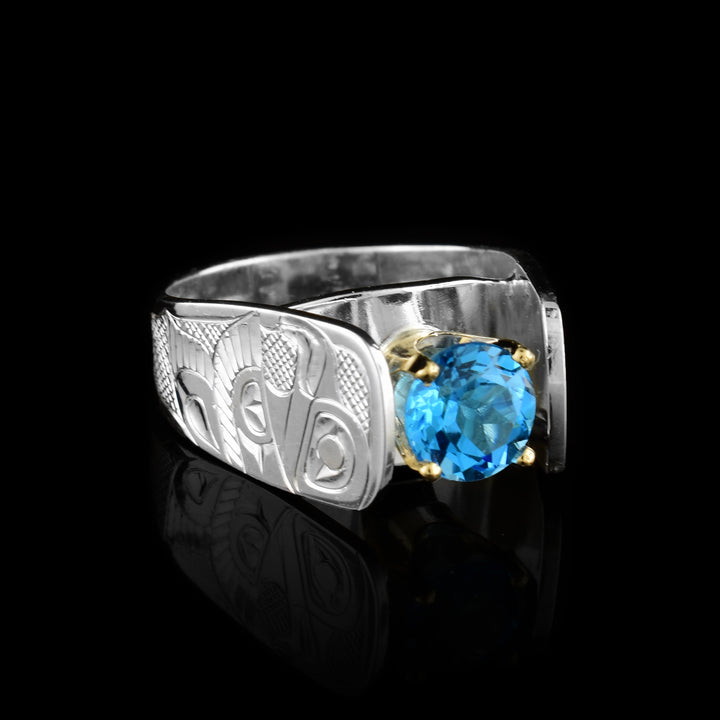This blue topaz solitaire ring has a wide and tapered band that extends in cathedral style above the blue topaz gem that is set in 14K gold claws. There are carvings made on the band that depict the Eagle. The Eagle is facing side ways at a side profile and has its wing extended out with feather details. Its eye, beak and tail are detailed and there are cross hatching designs in the background of the Scene.