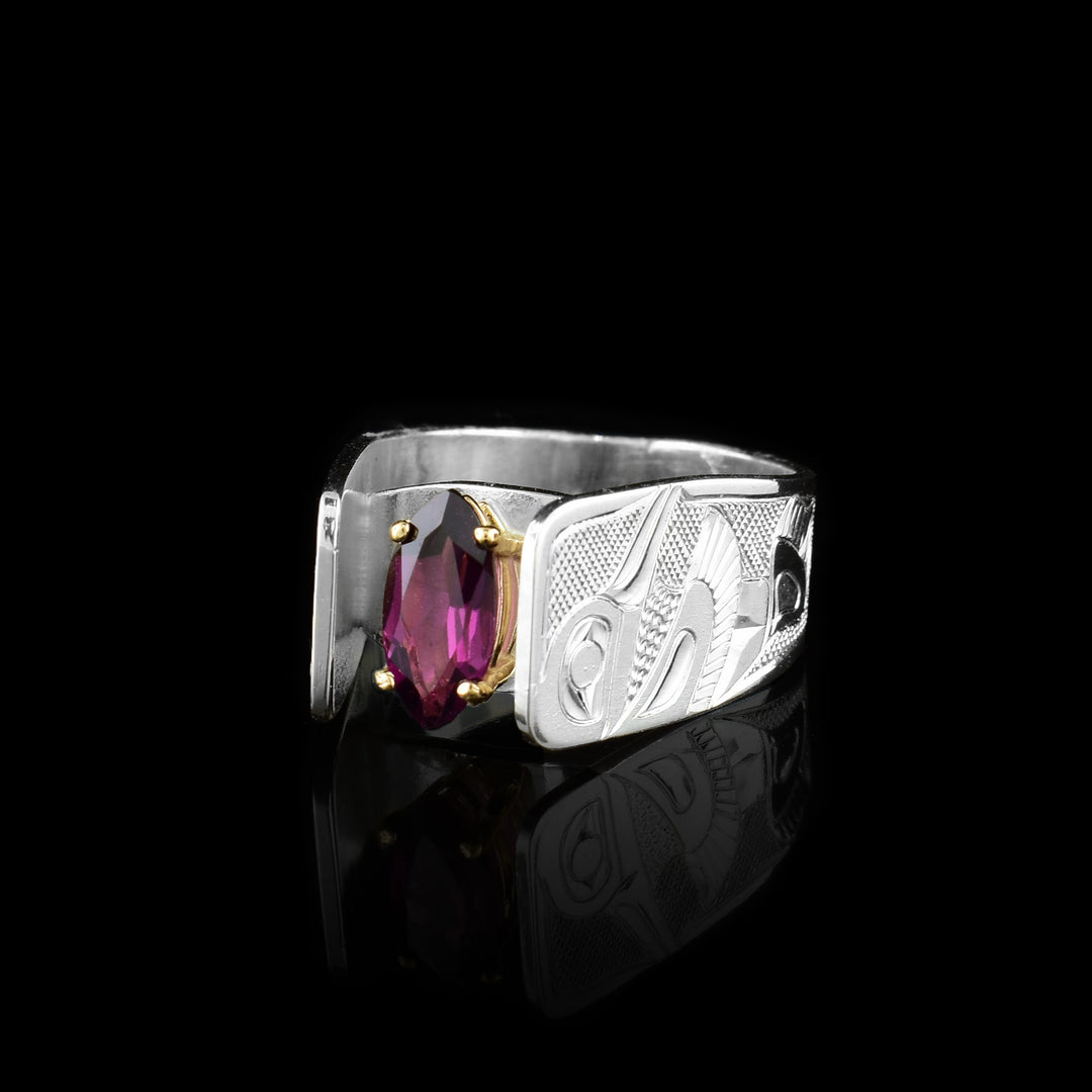 This wide Hummingbird ring has been hand carved by Tlingit artist Fred Myra. There are full body depictions of the Hummingbird legend carved into the sterling silver band of this marquis cut garnet solitaire ring.