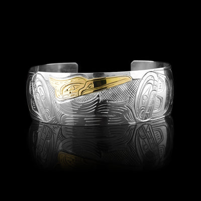 This silver and gold cuff bracelet has a single, wide band with a gap in the back. The band is sterling silver and the Blue Heron is carved into the center of the band. It is depicted in a side view and has a long neck and beak. Its head is done in 14K gold and its wings are depicted on both ends of the piece. The wings have feather details.