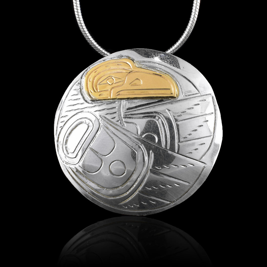 This gold and silver pendant has a large, circle shape. The Raven is depicted in the Piece with its head in the top and its two wings extending around it. The details of the ravens head and wings are included. The head of the raven is carved in 14K gold.
