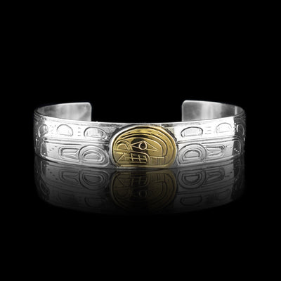This sterling silver and gold bangle bracelet has a single, thin band with carvings on the surface of it depicting the Orca. The head of the legend is carved into a 14K gold oval. On either side of the band, the body of the Orca with a dorsal fin and tail are carved.