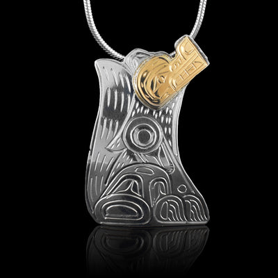This sterling silver and 14K gold Wolf pendant is shaped like the side profile of the Wolf. The Wolf's head is facing upward and its tail comes up to its head. The details of the wolf, including its fur, is carved into the surface of the pendant. The face of the legend has been made from gold.