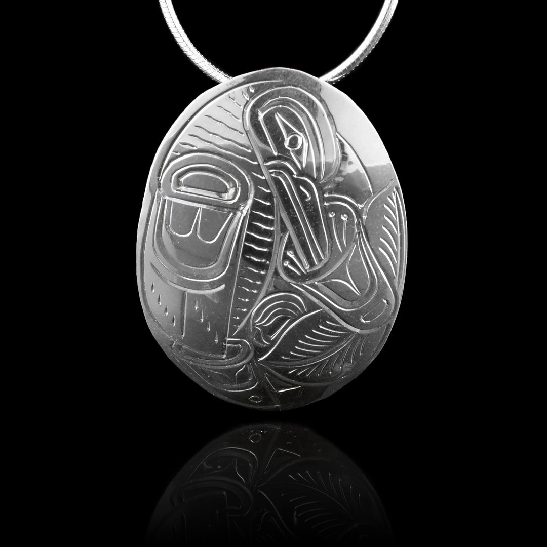 This Raven pendant has a large, oval shape, made from sterling silver and is longer than it is wide. The Raven is depicted in the side on the piece with its head facing down and its wing below it. On the other side is a flower with leaves around it and the Raven has its beak in the flower.