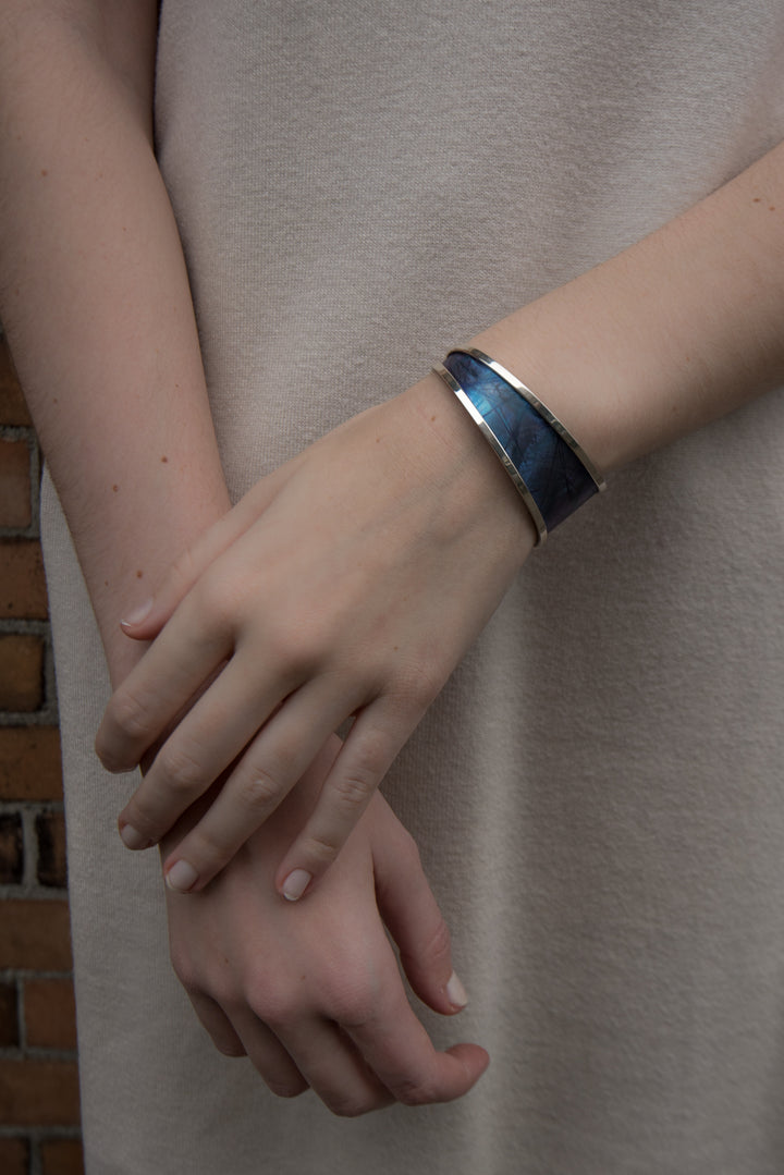 This titanium bracelet cuff is made from sterling silver and anodized titanium. It has a single band that tapers from 1" to 1/8" with a gap in the back. The band is made form titanium and is blue with designs achieved by anodizing the metal. There are silver accents surround the band.