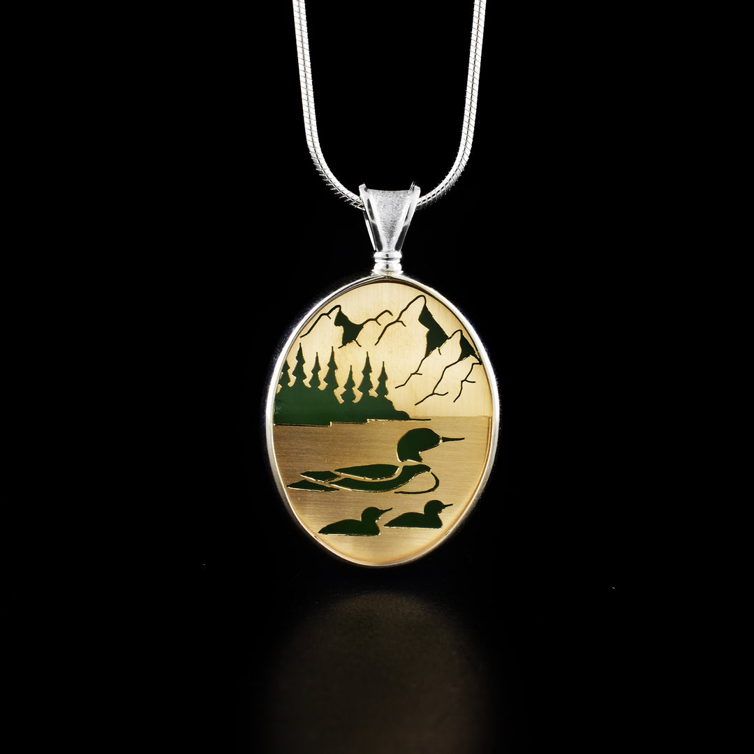 This bc jade pendant features a wildlife setting carved on 14k gold in which a mother loon and two her two babies are floating on the river with mountains and trees in the background.