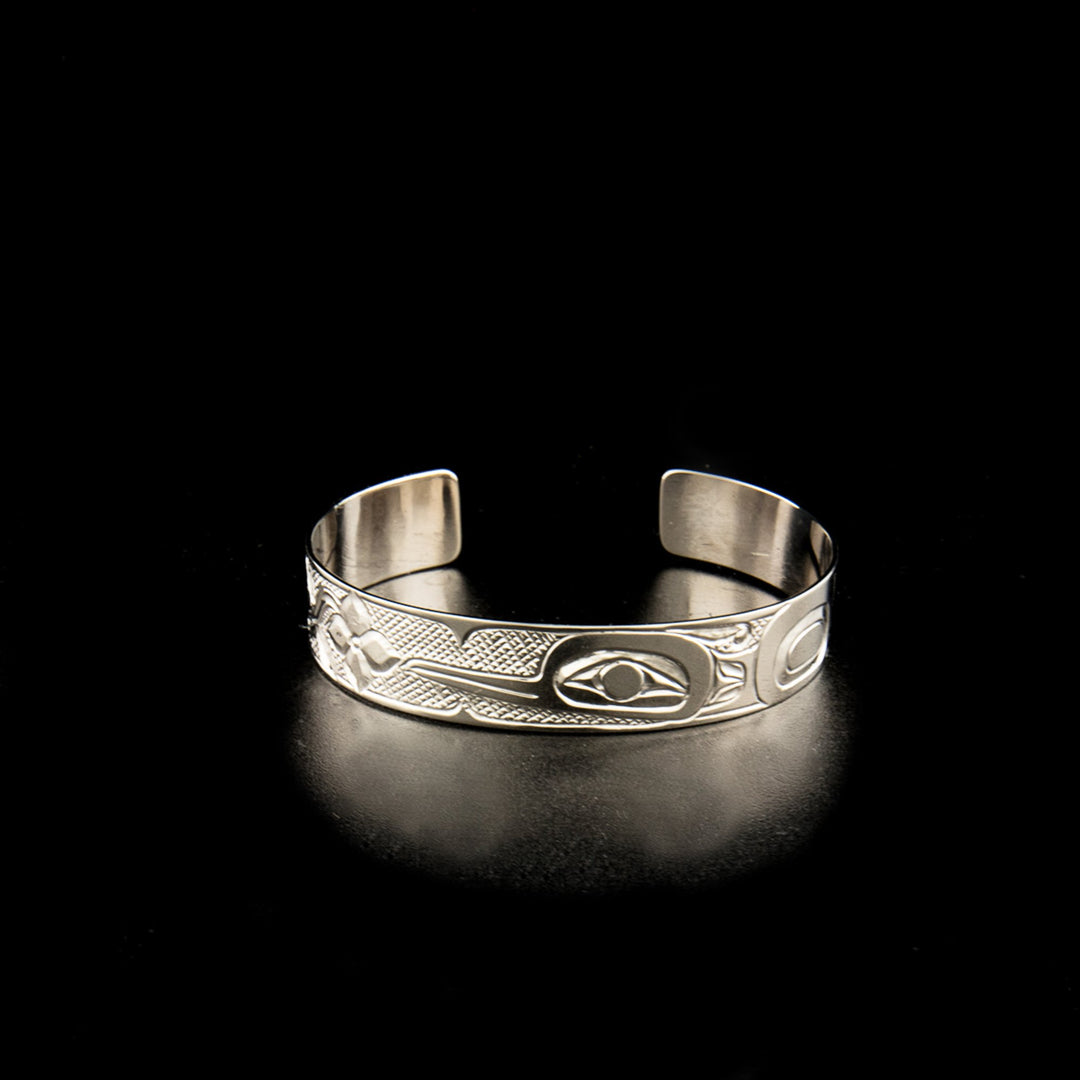 This 14K white gold cuff bracelet has a single, thin band with a gap in the back. There is a side profile depiction of the hummingbird carved into the band. Its beak is facing a small flower and its ye is large. Its wings can be seen tucked behind it and there are cross hatching designs in the background of the scene.