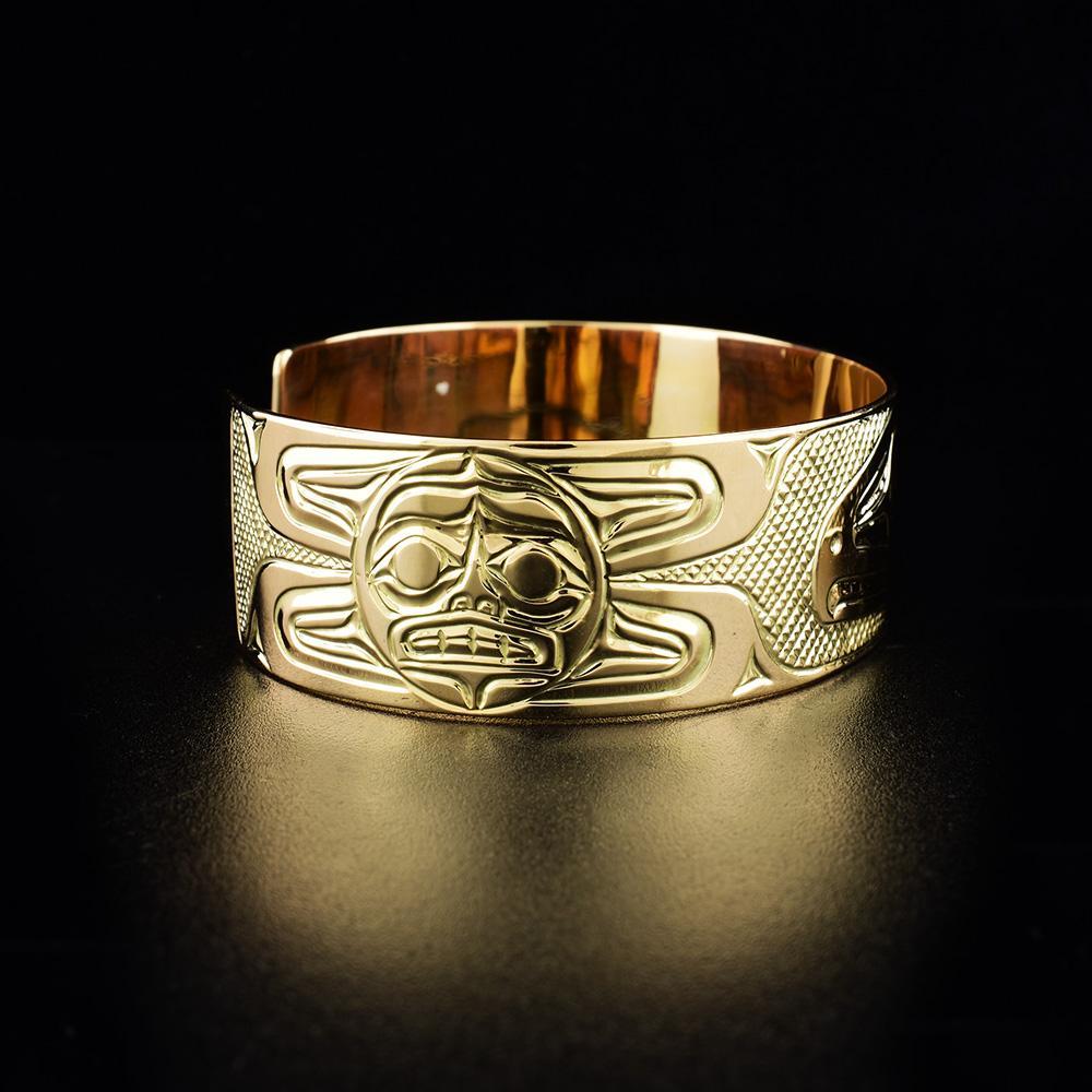 This 14K gold cuff bracelet has a single, wide band with a gap in the back. There are depictions of the Sun and the Raven carved into the surface of the band.