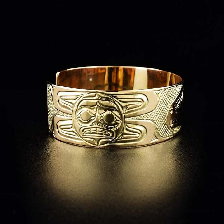 This 14K gold cuff bracelet has a single, wide band with a gap in the back. There are depictions of the Sun and the Raven carved into the surface of the band.