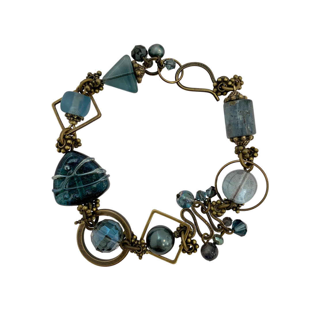 This blue crystal bracelet is made from Austrian Crystal, Brass, Freshwater Pearls, BC Artist Studio Glass, Recycled Glass, Kyanite, and Iolite. The chain bracelet features beads of various shape and material.