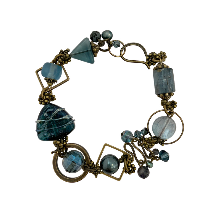 This blue crystal bracelet is made from Austrian Crystal, Brass, Freshwater Pearls, BC Artist Studio Glass, Recycled Glass, Kyanite, and Iolite. The chain bracelet features beads of various shape and material.