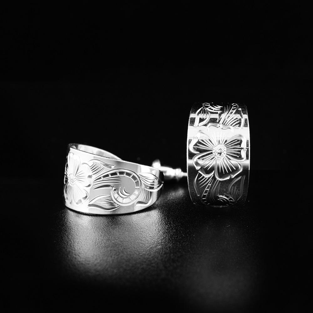 These silver flower earrings are made from sterling silver and have a single wide band curved into a hoop with a gap in the back. There are floral designs carved into the surface of the earrings.