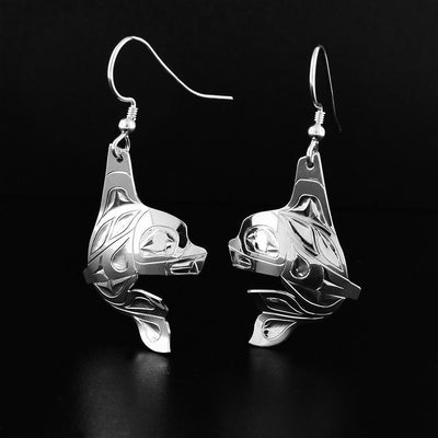 Sterling Silver Dancing Orca Earrings by Fred Myra. Each earring is in the shape of an orca facing inward. The artist has hand-carved intricate designs along the body of the orca that represent the orca's black and white spots. The orca has a large dorsal fin to which the hook is connected to.