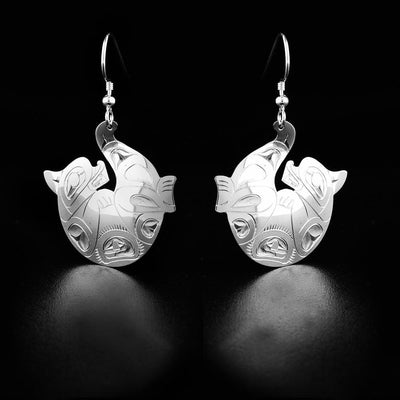 Sterling Silver Otter Earrings by Fred Myra. Each earring is in the shape of an otter laying on their back. The artist has hand-carved intricate designs along the otter's body to represent the arms and legs and the fur of the creature. The tail of the otter curves upward and that is where the hook of each earring is attached.