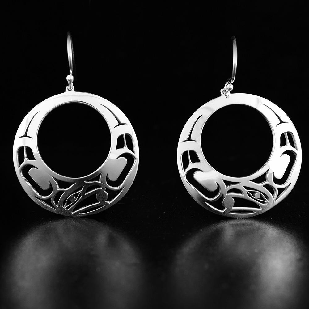 These cutout, circle dangle earrings are made from sterling silver and have circular hangs with a large, circle cutout. There are intricate cutout designs that depict the Raven on the surface of the hangs.