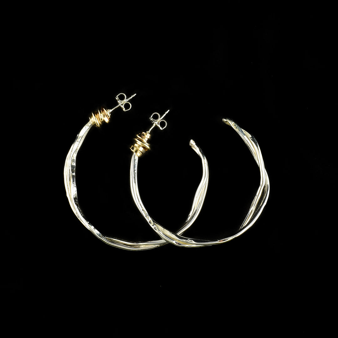 Sterling Silver Twist of Fate large hoop earrings handcrafted by artist Joy Annett. There are two sterling silver wires twisted around each other and curved into hoops. There are 14K gold filled wrappings below the stud hooks accenting the piece.