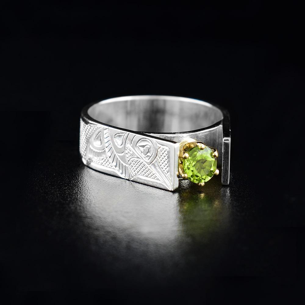 This Hummingbird cathedral ring has a wide, sterling silver band and a peridot gem mounted in the center. The Hummingbird is carved in a side profile with its head looking up towards the gem and its wing at its side. The band extends up and frames the peridot ring which is claw mounted in 14K gold.