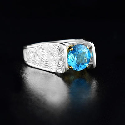 Solitaire Orca Ring with Blue Topaz by Fred Myra. The artist has hand-carved the profile of two full-bodied orcas on either side of the blue topaz stone he has set in the center of the ring. The background of the ring has been hand-carved into a neat crisscross pattern to allow for the orca legends to stand out. The blue topaz has been set using a 14k gold claw setting. The ring tapers down at the back.