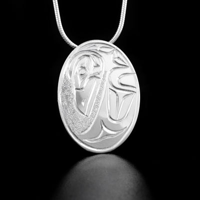Sterling Silver Oval Raven Pendant by Paddy Seaweed. The design depicts the profile of a raven's head facing downward with a large, spread out wing underneath the head. The background has been delicately hand carved to allow for the image of the raven to pop out.