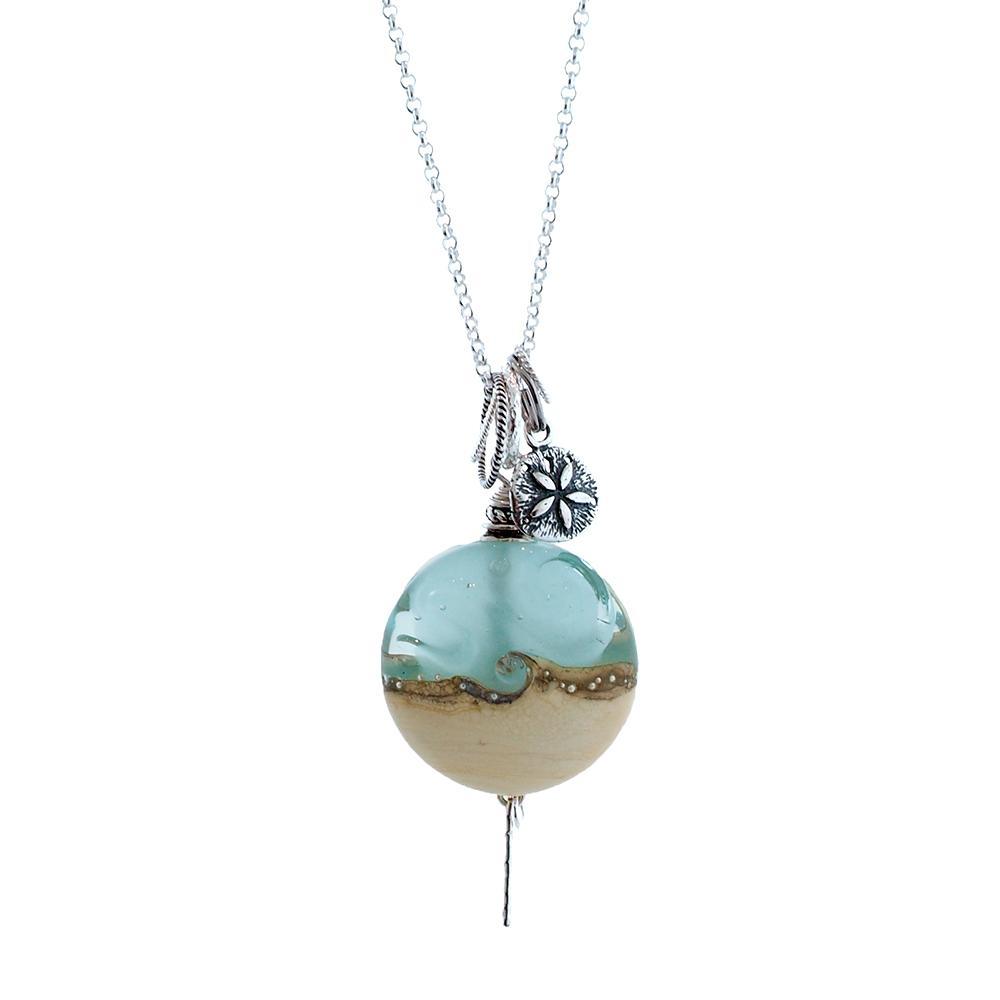This glass pendant necklace is made from lamp worked glass and sterling silver. The pendant has a glass bead that depicts the sand and the sea, and above the pendant is a silver sand dollar charm.
