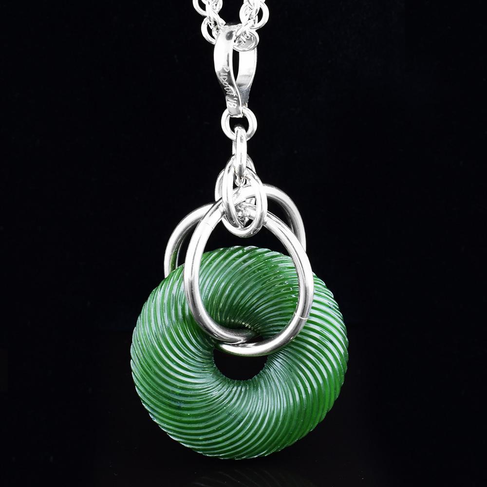 This BC jade pendant necklace has a large, disc shaped jade piece with multiple sterling silver hoops linked through it. The jade piece has a ribbed texture on the surface of it.